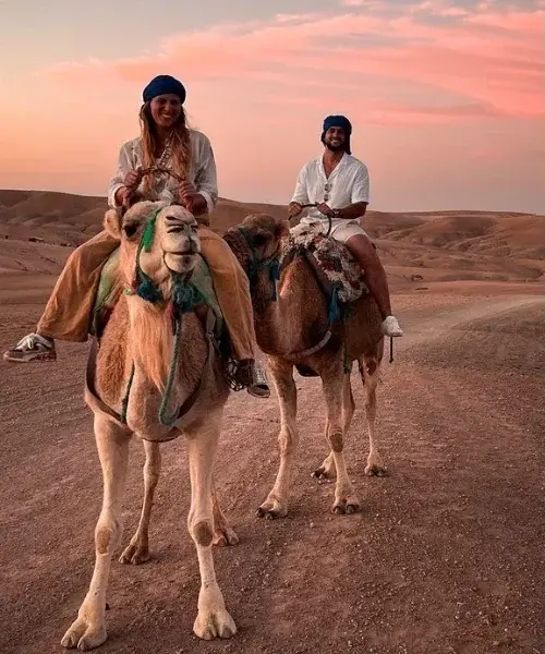 Two travellers on camels crossing the Agafay Desert under a pink evening sky