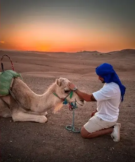 Orange sunset over the rolling hills of the Agafay Desert, with a resting camel and its handler in the foreground