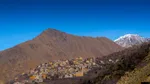 Imlil village spread across a sunlit hillside beneath dry brown peaks, with a snow-capped summit visible in the distance