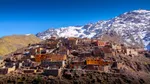 Colorful Imlil village of ochre and terracotta houses clinging to a rocky mountainside, with a snow-capped High Atlas peak towering behind