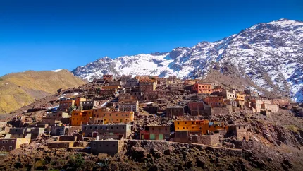 Colorful Imlil village of ochre and terracotta houses clinging to a rocky mountainside, with a snow-capped High Atlas peak towering behind