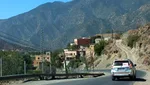 Mountain road through an Ouirgane village, with a car driving past hillside houses below the High Atlas