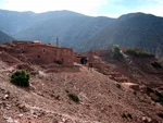 Traditional red mudbrick kasbah-style buildings on a rocky Ouirgane hillside, backed by pine-forested mountains